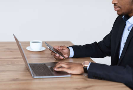 Cropped of black man in suit businessman working in office, using gadgets and drinking coffee, side view, copy space. African american manager typing on laptop and holding mobile phoneの写真素材