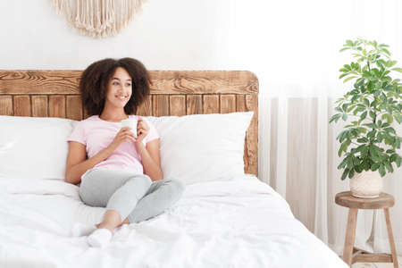 Weekend, good morning, positive mood and vitality. Happy african american female sitting on bed, holding cup of hot drink and enjoying morning in cozy comfort modern bedroom interior, free spaceの写真素材