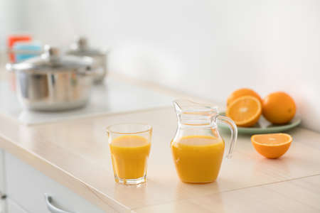 Proper nutrition, vitamins, morning breakfast and healthy food. Fruits on plate, glass and jug of orange juice on furniture, next to stove with pans, white walls in kitchen, in daylight, copy spaceの写真素材