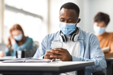 Chatting With Friends. Portrait of african american male student wearing disposable surgical mask, using mobile phone and browsing social media at class during lecture, sitting at table in universityの写真素材