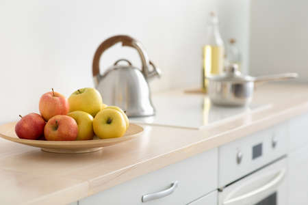 Modern kitchen and healthy eating at home, minimalist cozy interior in morning. Colorful apples on plate, on furniture, next to stove with metal kettle, utensils, white walls, in daylight, copy spaceの写真素材