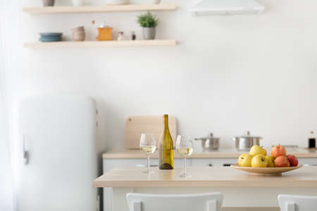 Classic scandinavian kitchen with wood and white details, minimalist interior design, rent. Plate with apples, glasses and bottle with light wine and utensils on table, refrigerator and light wallsの写真素材