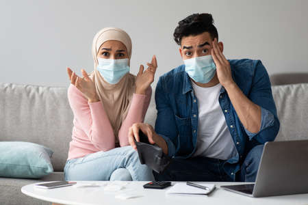 Worried muslim couple in protective face masks sitting on couch with empty wallet in front of laptop...