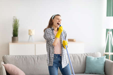 House cleaning is fun. Cheerful young lady using mop as mic, singing song while doing cleanup at living room. Millennial housewife pretending to be rock star while doing domestic dutiesの写真素材