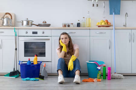 Exhausted young housewife sitting on floor at kitchen, surrounded by cleaning supplies, tired from housework. Overworked woman needing break from domestic duties, cannot stand doing house choresの写真素材