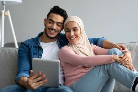 Happy middle-eastern young couple sitting on sofa at home and looking at digital tablet screen,...