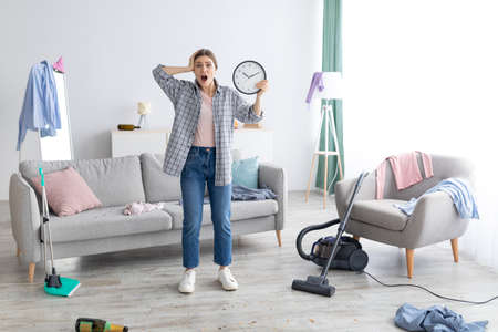 Shocked young lady with clock grabbing her head in terror, standing in messy room after party, too late to clean apartment before return of her parents. Space for textの写真素材