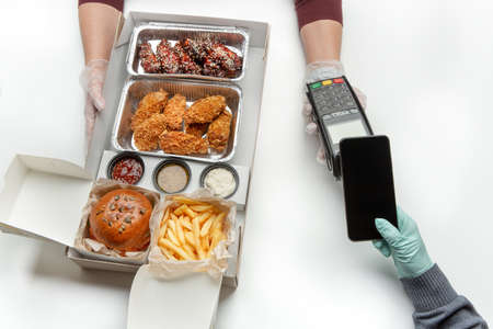 Junk food and home delivery during quarantine. Hands of man in rubber gloves hold phone with empty screen, male pays at terminal, courier gives order with fried meal dishes on white tableの写真素材