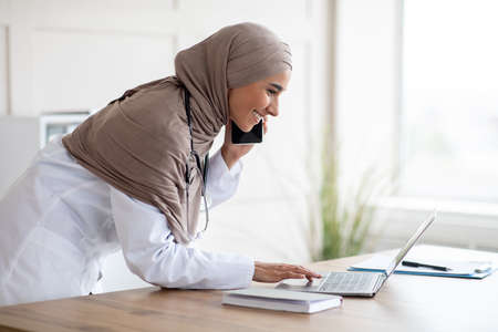 Side view of female doctor young beautiful lady in headscarf arranging appointment on mobile phone, having conversation with patient and looking at her schedule on laptop, clinic interior, copy spaceの写真素材