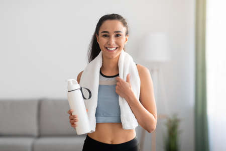 Cheerful brunette woman in sportswear having break after training at home, holding bottle with water...