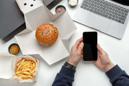 Food delivery to workplace, lunch for employee in office. Burger in cardboard box, french fries and sauce in container, laptop, hands of male worker manager hold phone with blank screen on white tableの写真素材