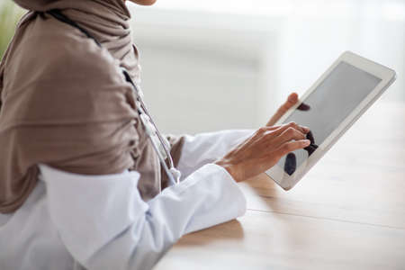 Cropped of female doctor in hijab using gadgets at workplace, holding digital tablet. Unrecognizable therapist muslim woman having online conversation with her patients, using pad, side viewの写真素材
