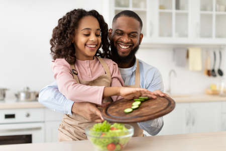 Making Healthy Meal. Portrait of happy african american dad teaching his little girl how to prepare vegetarian salad, smiling father and cute daughter cooking together at home.の写真素材
