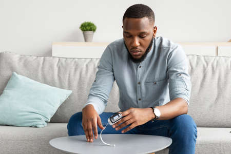 African Guy With Pulse Oximeter Clip On Hand Measuring Level Of Oxygen Saturation Sitting On Couch At Home. Ox Measurement Device, Pulseoxymetry Machine Concept. Selective Focusの写真素材