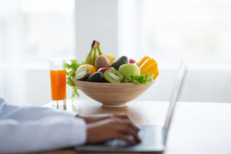 Dietology and healthy nutrition concept. Weight loss consultant working on laptop, giving online consultation to patient at clinic, focus on bowl of fruits and veggies with glass of juiceの写真素材