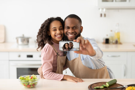 Spending Time Together. Portrait of happy african american family taking selfie while cooking in the kitchen at home, using mobile phone. Smiling black dad and daughter making photoの写真素材