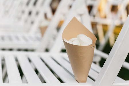 Celebrate of marriage, accessories for boho wedding ceremony outside. White rose petals in paper bag on guest chair for showering newlyweds on green lawn in summer day, flat lay, close up, free spaceの写真素材