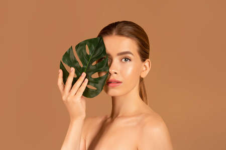 Portrait of pretty young woman with smooth skin and nude makeup closing her eye with monstera leaf on brown studio background. Attractive lady hiding behind tropical plant. Skincare and beauty conceptの写真素材