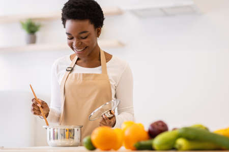 Happy African Woman Cooking Dinner In A Pot Standing In Modern Kitchen At Home, Wearing Apron. Black Housewife Preparing Meal Indoors. Healthy Food Recipes, Household And Nutritionの写真素材
