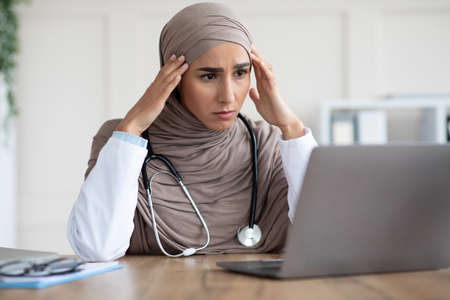 Worried woman doctor beautiful woman in hijab having headache while working with laptop, looking at screen and touching her temples, having difficulties at job, closeup, copy space, clinic interiorの写真素材