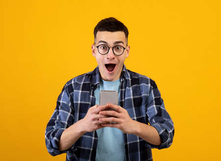 Handsome young guy in glasses looking at his smartphone in shock on orange studio background....
