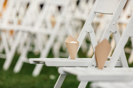 Preparation for wedding ceremony, for shower with petals of newlyweds outdoor. White folding chairs with paper packages on green lawn for marriage rite in summer, selective focus, flat lay, free spaceの写真素材