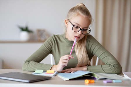 Concentrated school girl in glasses teenager doing homework in her cozy room, looking at school book and taking notes, copy space. Smart caucasian teen girl studying at home, kids education conceptの写真素材