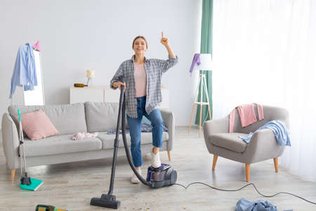 Happy young lady with vacuum cleaner having cleanup idea at messy room after party. Millennial female student hoovering dirty apartment, pointing finger up, gesturing eurekaの写真素材