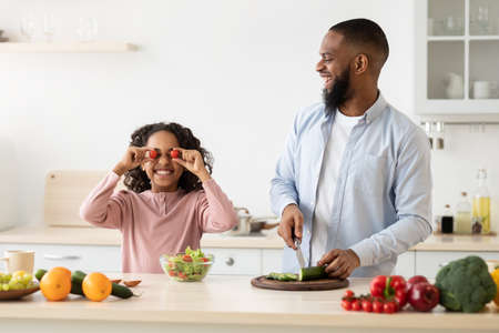 Kitchen Fun. Portrait of funny black little girl playing with food, pretending tomatoes vegetables as glasses having fun in the kitchen, cooking meal together with smiling father cutting cucumberの写真素材