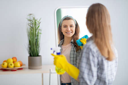 Cheerful millennial housewife wiping mirror with rag, using spray detergent to clean looking glass at home. Young woman doing domestic chores, keeping her apartment tidyの写真素材