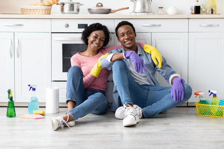 Tired black couple in rubber gloves sitting on floor next to stove among house-keeping supplies in cozy kitchen, hugging, resting after cleaning apartment, smiling at camera, copy spaceの写真素材