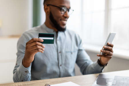 Mobile Banking Application. Black Businessman Using Credit Card And Smartphone At Workplace Sitting In Modern Office. Shopping App For Mobile Phone, E-Commerce Concept. Selective Focus, Croppedの写真素材