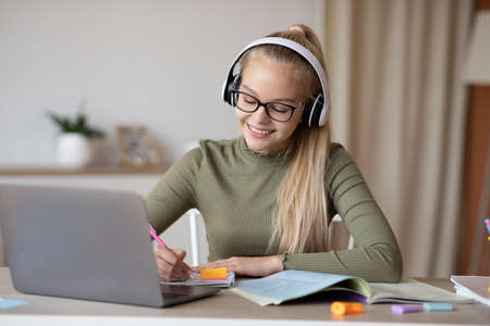 Smiling teen girl in wireless headset having online lesson while staying home during coronavirus...