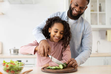 Togetherness. Portrait of smiling girl learning how to cook and cut vegetables, positive black dad helping his daughter slicing cucumber with knife, making tasty delicious dinner together in kitchenの写真素材
