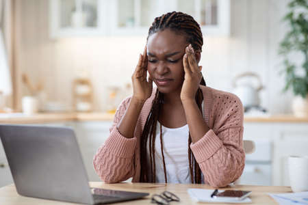 Migraine Concept. Stressed African American Woman Suffering From Headache While Working On Laptop At Home, Upset Lady Sitting At Desk With Computer In Kitchen And Touching Temples, Free Spaceの写真素材