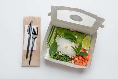 Modern salad and useful food, delivery and health care. Tasty asian dish with fish and vegetables in box, fork, knife and napkin on eco paper bag, isolated on white background, top view, copy spaceの写真素材