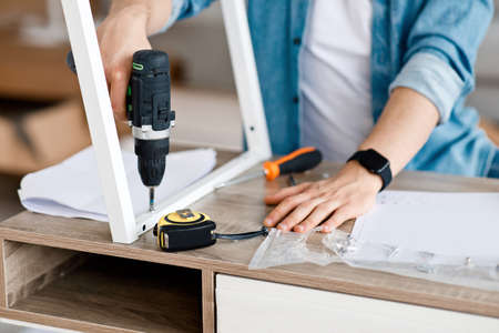 Man assembles table and furniture repair home master. Millennial male hand with drill screwing...