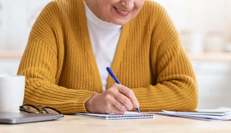 Cropped of old woman sitting at table with papers, laptop and mobile phone in kitchen, taking notes,...
