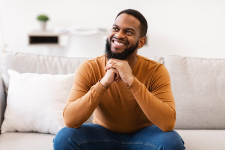 Dreamy Black Guy Thinking Smiling And Sitting On Couch At Home. African American Man Dreaming About Success Looking Aside Posing Indoors Conceptの写真素材