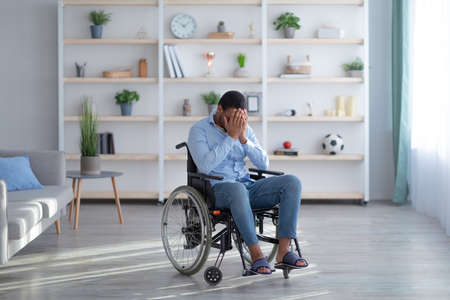 Depression and loneliness in disabled adults. Young black guy in wheelchair crying at living room,...