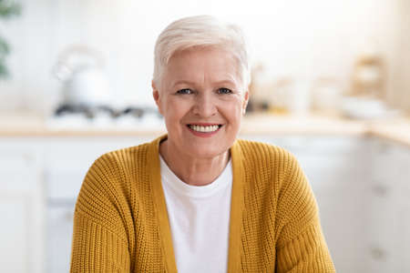 Portrait of happy senior lady sitting in kitchen at home, closeup, copy space. Attractive old woman...