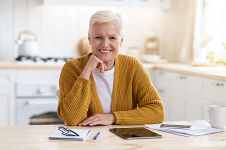 Happy senior woman freelancer working from home, sitting in kitchen, using digital tablet and...