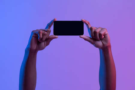Black Male Hands Holding Smartphone With Black Screen In Horizontal Orientation Under Vivid Neon Light Over Purple Background, Mockup Image With Copy Space For Your App, Game Or Website Designの写真素材