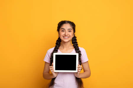 Great Application. Smiling Indian Lady Showing Tablet Computer With Empty Screen To Camera Recommending New App Posing Standing Over Yellow Studio Background. Digital Tablet Display Mockup For Advertの写真素材
