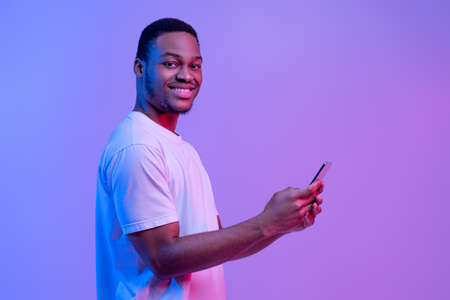 Happy Smiling Black Man Using Smartphone While Standing Under Neon Light Over Purple Background, Millennial African American Guy Enjoying Modern Mobile Technologies, Looking At Camera, Copy Spaceの写真素材