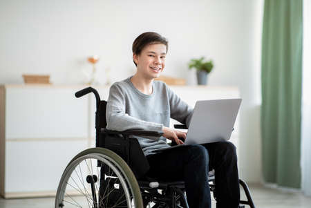 Happy disabled teenager in wheelchair using laptop computer for online studies at home. Positive handicapped teen boy communicating with teacher remotely, having webinar indoorsの写真素材