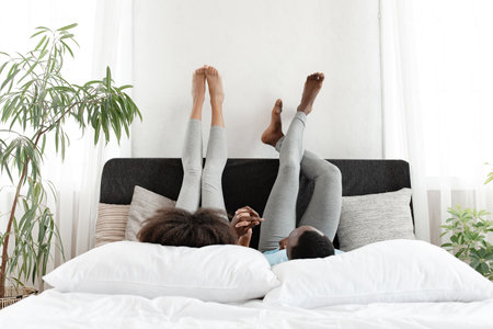 Communication in free time and relaxing on bed, having fun together and lazy at home. Happy millennial african american lady and man lie with raised legs in bedroom interior in morning, copy spaceの写真素材