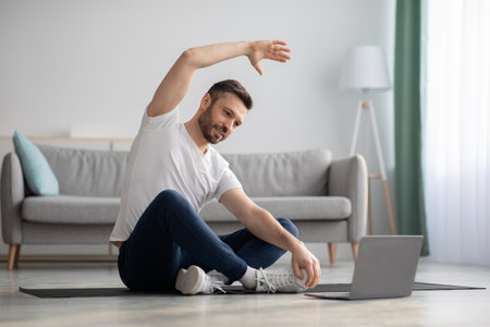 Positive athletic bearded middle-aged man stretching at home, using laptop, watching sport videos on Internet, having fitness class online, copy space. Healthy lifestyle, sport on self-isolationの写真素材