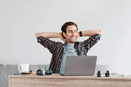 Break at work at home, repairman, handyman or blogger enjoying free time. Smiling young guy looks at empty space, sits at table with laptop, in living room with cup and tools for assembly furnitureの写真素材