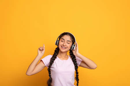 Happy young indian woman in wireless headset listening to music with closed eyes, calm millennial lady enjoying good sound with modern headphones, isolated on yellow studio backgroundの写真素材